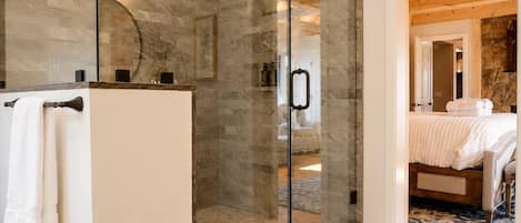 Un baño moderno con ducha envuelta en vidrio, un lavabo blanco y una habitación visible con cama y suelo de madera.