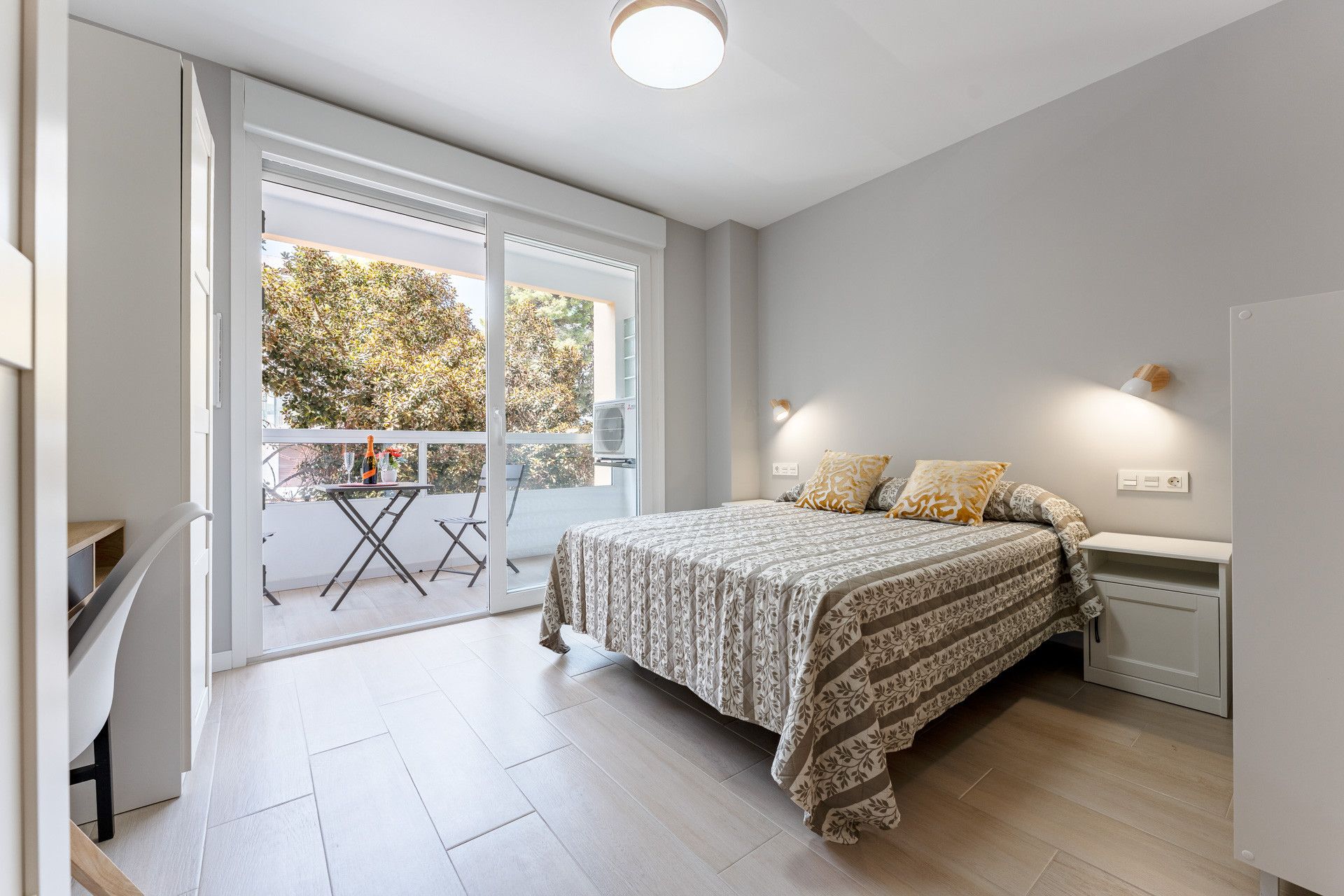 Une chambre à coucher comprenant un lit, des tables de chevet, une commode, une fenêtre donnant sur des arbres et un balcon avec une table et des chaises.
