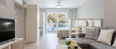 Una sala moderna con televisor de pantalla plana, un sofá gris, una mesa de centro de madera con un florero y un balcón con mesa y sillas.