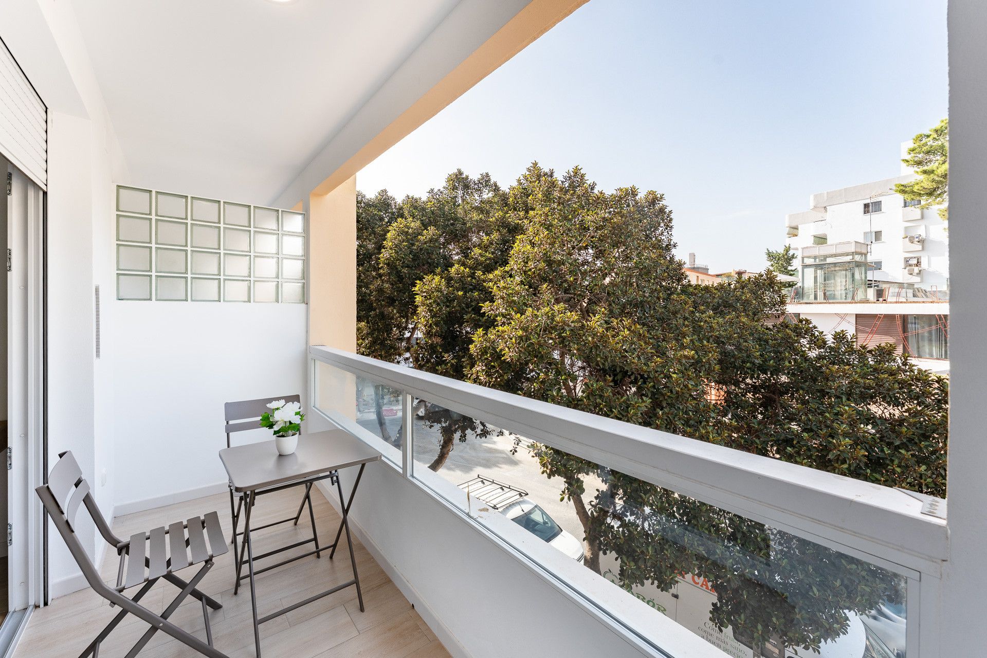 Un balcón con una mesa, sillas, una planta en maceta y vistas a árboles y edificios.
