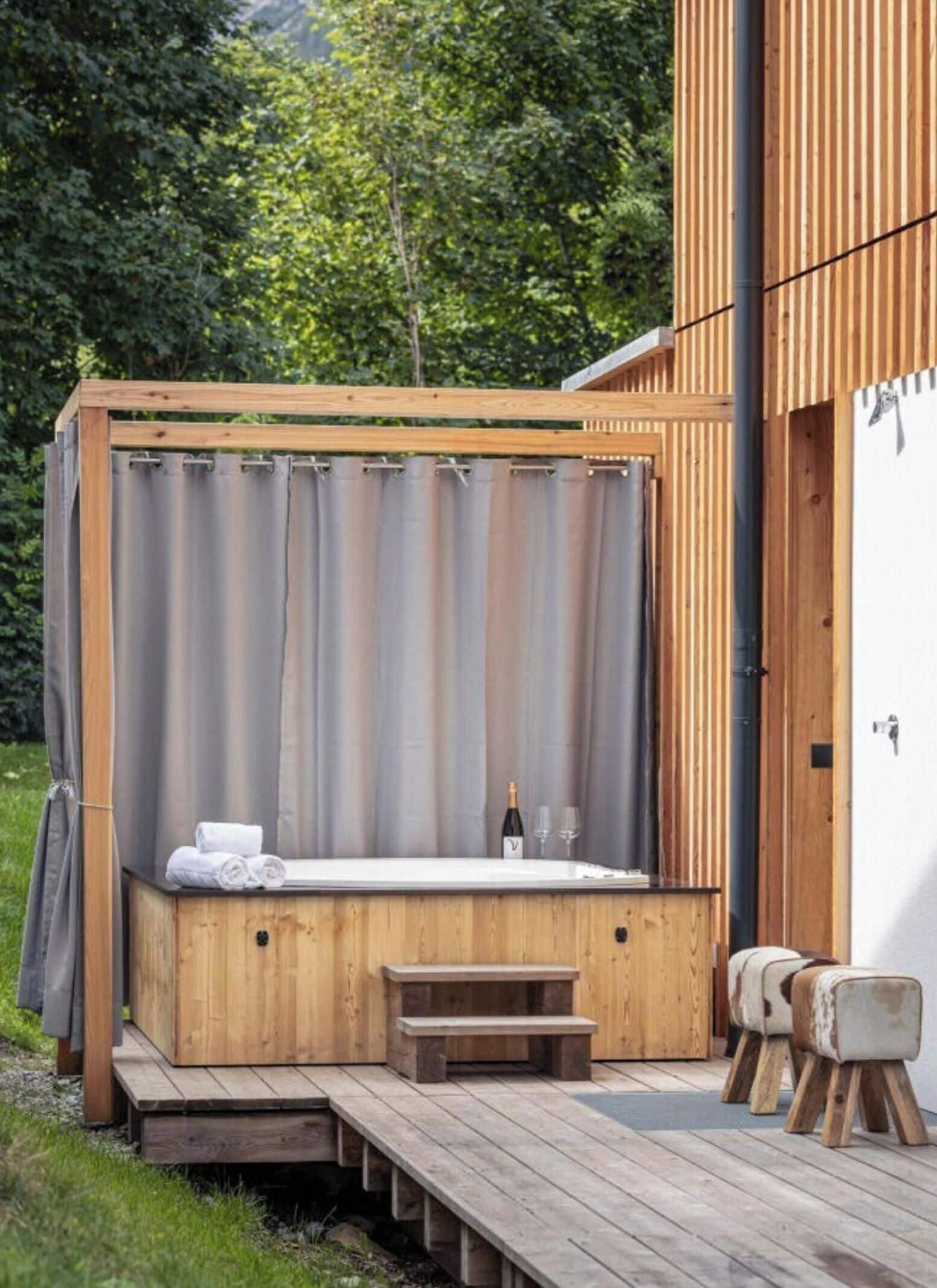 Un bain à remous extérieur en bois, doté d’un banc et de marches, entouré d’une terrasse en bois et d’un bâtiment blanc.