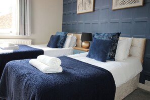 A hotel room with two beds, each with a blue bedspread and white linens. There are decorative pillows, a bedside lamp, and a small table with a lamp. - The Hampton (Scarborough)