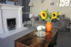 A living room with a fireplace, a coffee table with a vase of sunflowers and white flowers, and a grey sofa. - The Hampton (Scarborough)