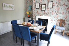A dining room with a wooden table set for four, blue upholstered chairs, a fireplace, and floral wallpaper. - The Hampton (Scarborough)