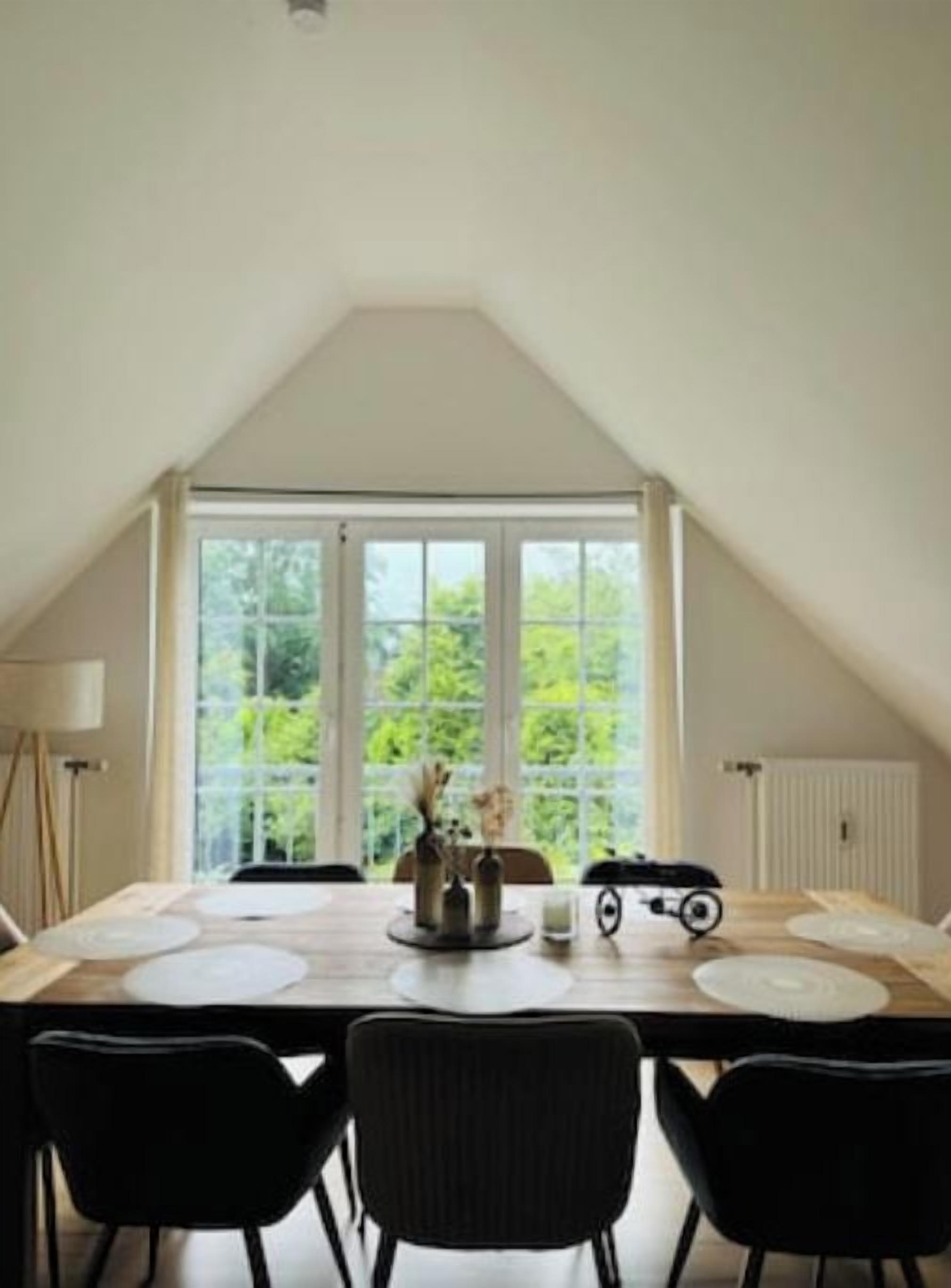 A dining area with a table set for four, a large window with a view of greenery, and a sloped ceiling.