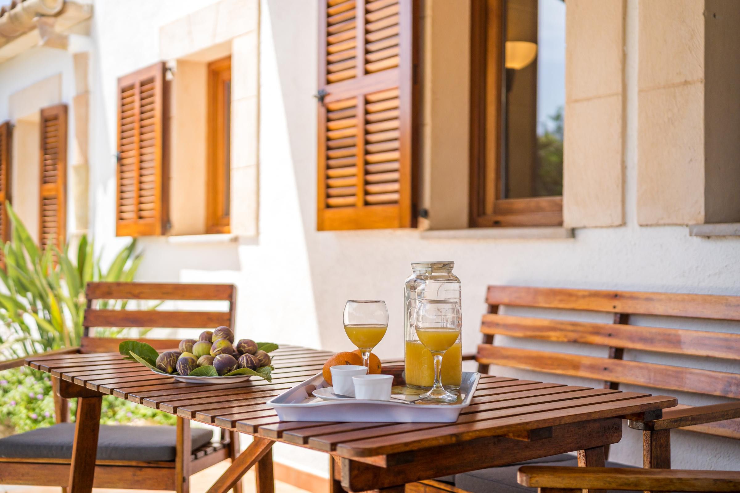 An outdoor wooden table with a tray containing fruits, a glass of juice, and a bottle of juice.