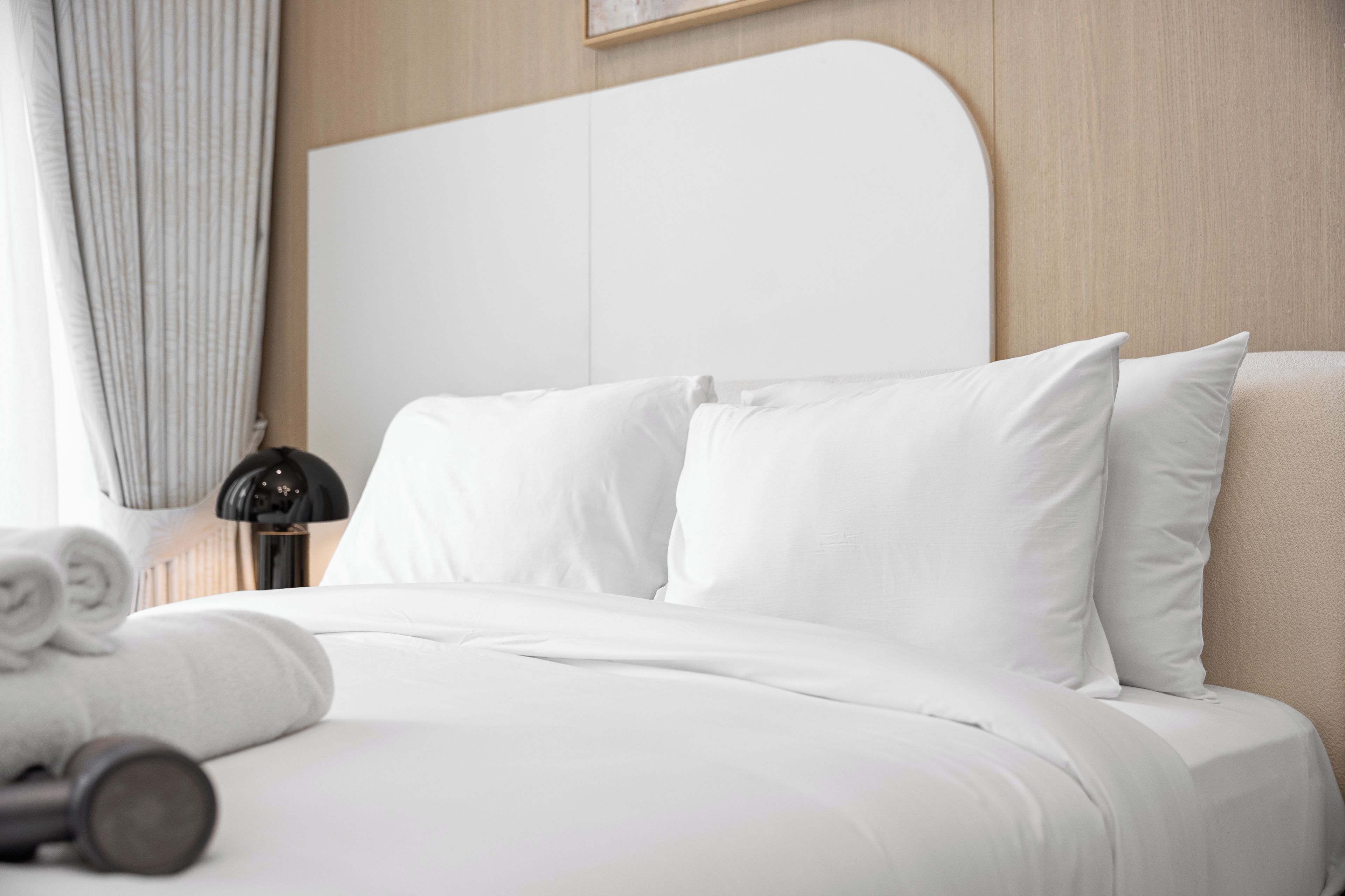 A neatly made bed with white linens, a black lamp, and rolled towels on the side.