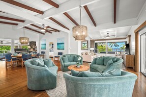 A spacious living area with teal armchairs, a wooden coffee table, and a large window offering a view of the outdoors. - Private Poipu Retreat with Rooftop Ocean Views, Hot Tub and Outdoor Living (Koloa)