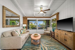 A living room with a sofa, coffee table, TV, and windows with a view. - Private Poipu Retreat with Rooftop Ocean Views, Hot Tub and Outdoor Living (Koloa)