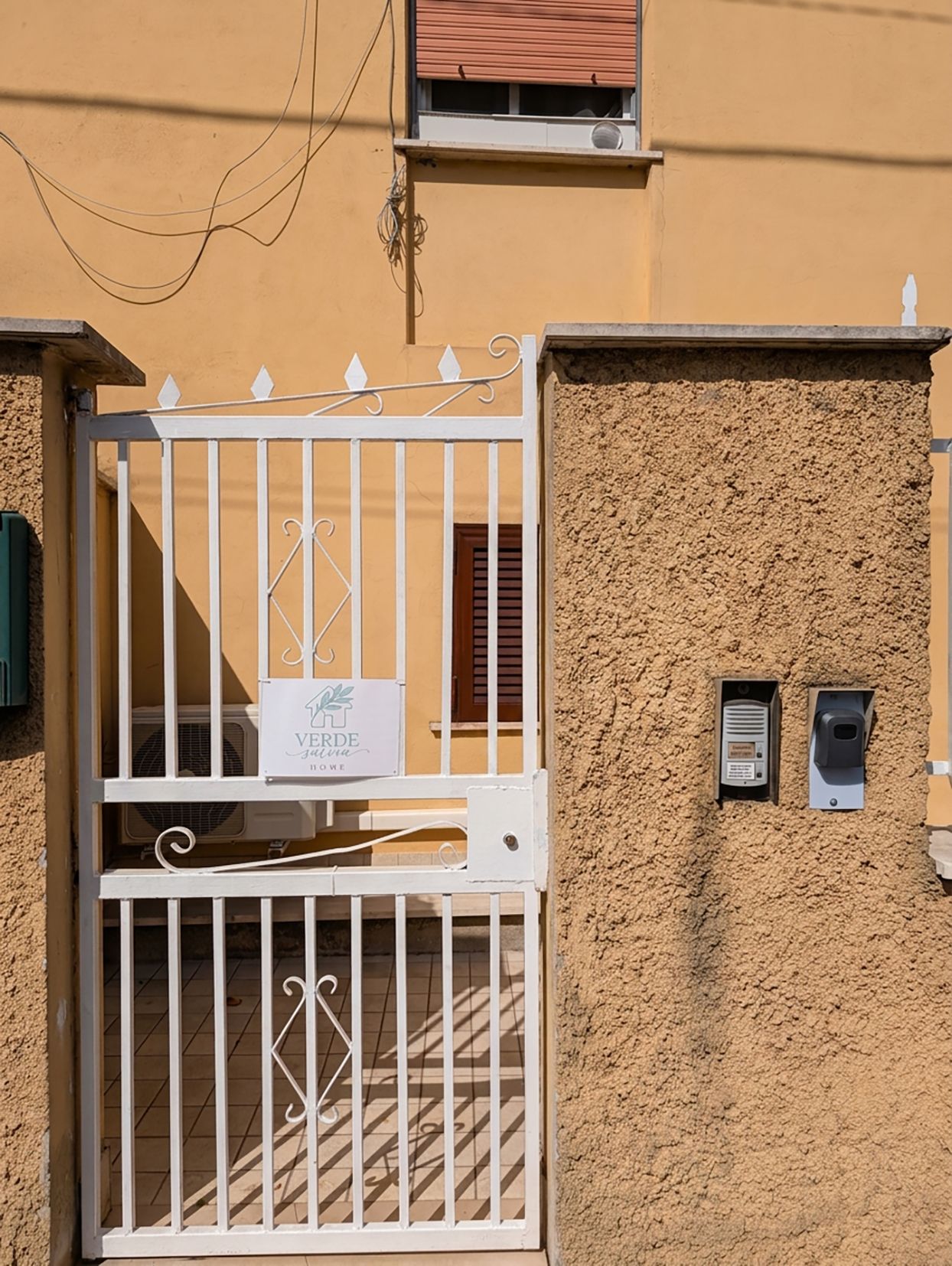 A gated entrance with a sign, a textured wall, and a small electronic device mounted on the wall.