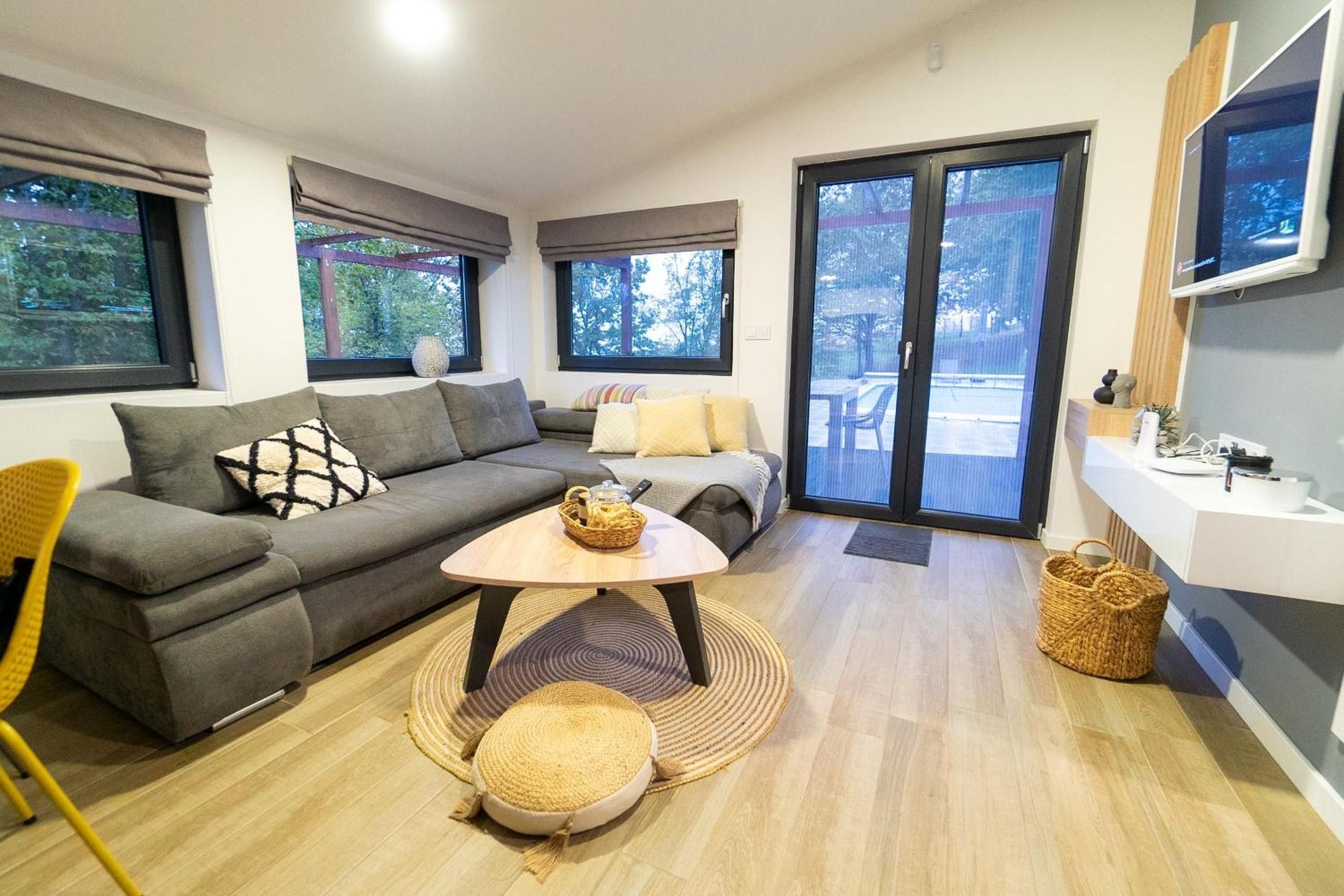 A modern living room with a grey sofa, a small round table, and a yellow chair. There is a large window with a view of trees and a balcony.