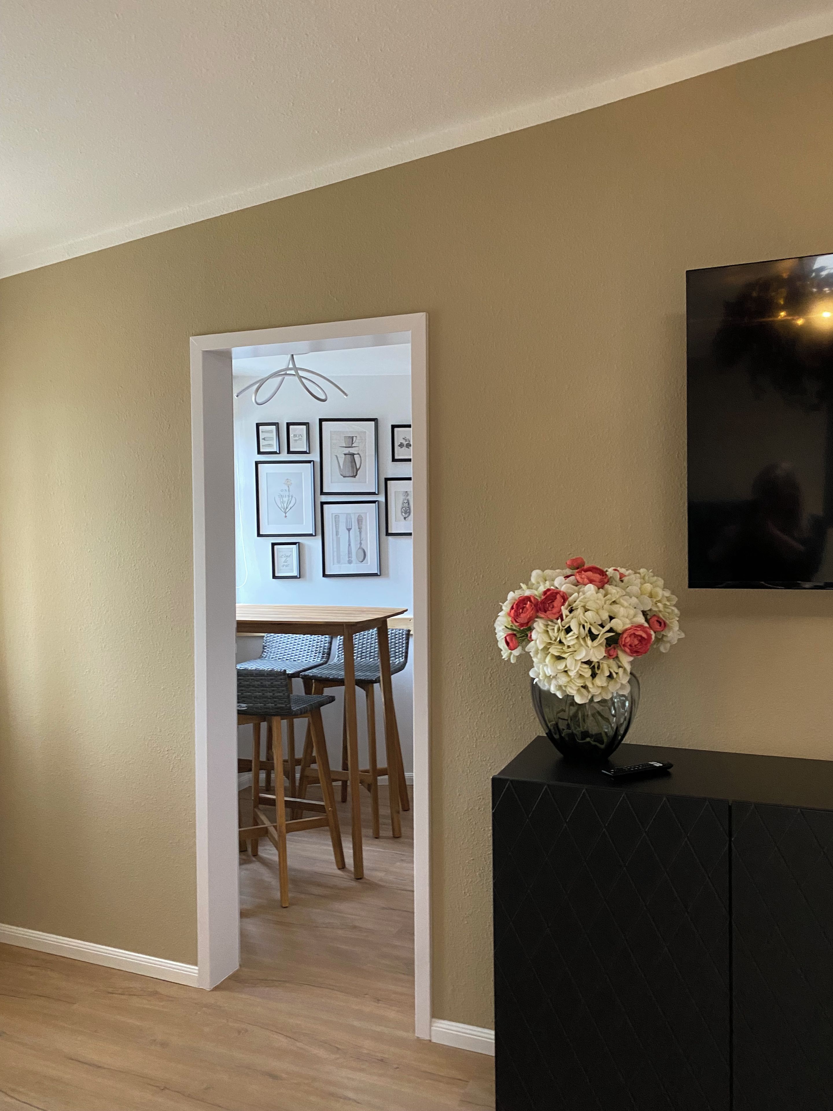 Quarto com área de jantar, armário preto com um vaso de flores e televisão fixada na parede.