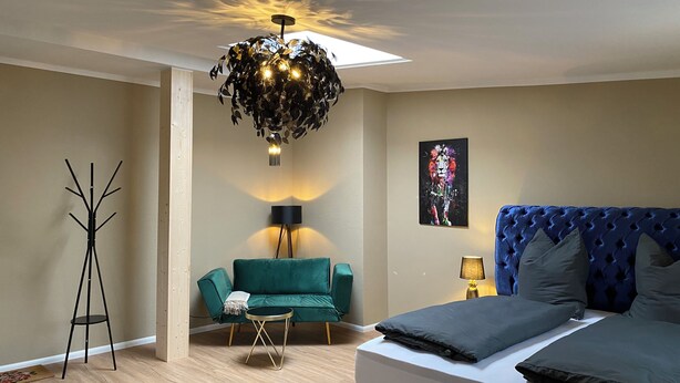 A bedroom with a bed, a green sofa, a standing lamp, and a ceiling light fixture. - Zur Post 1 (Bautzen)