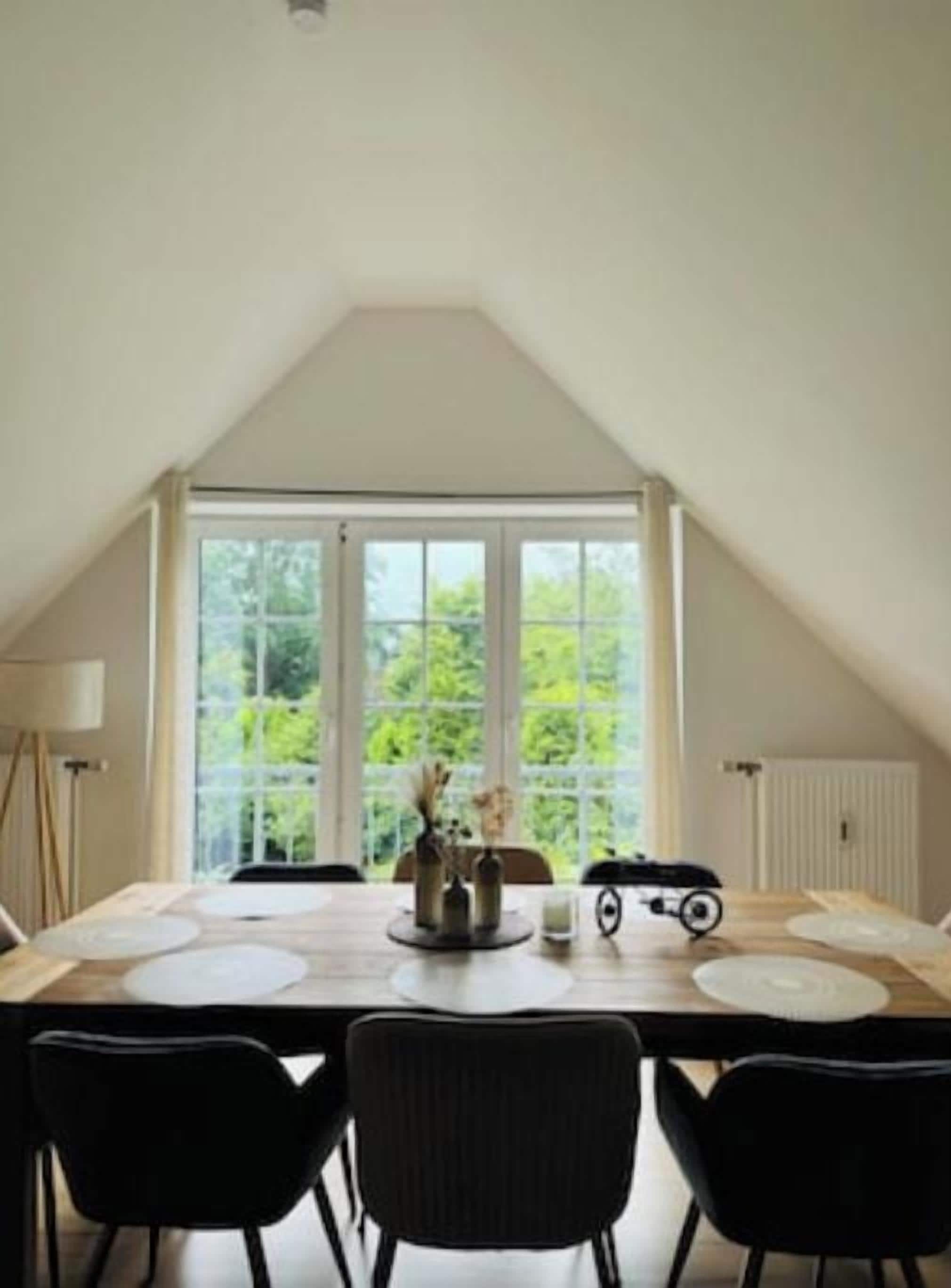 A dining area with a table set for four, a large window with a view of greenery, and a sloped ceiling.