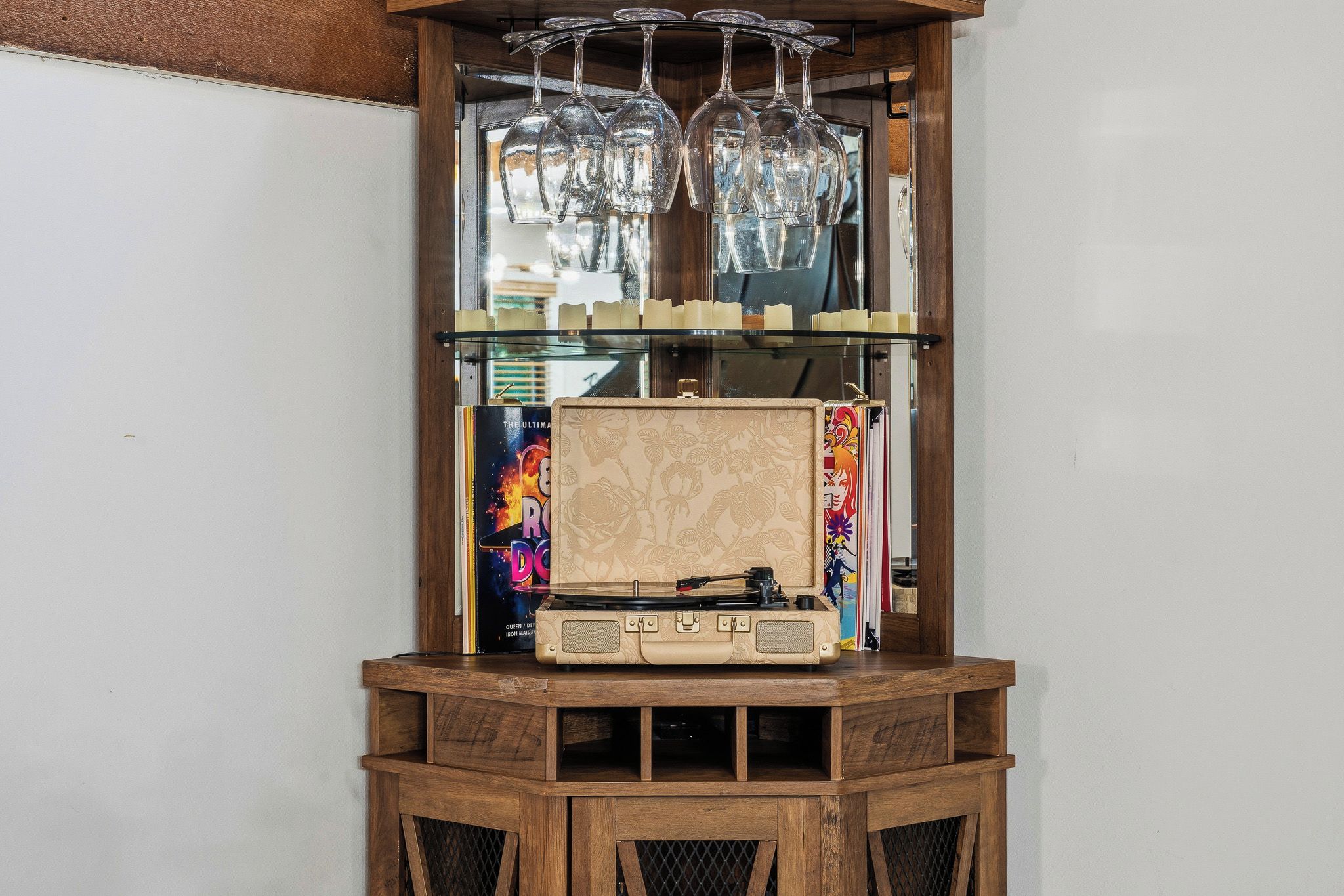 Un mueble de madera con un tocadiscos, una repisa de vidrio con copas de vino y un objeto decorativo con motivos florales.