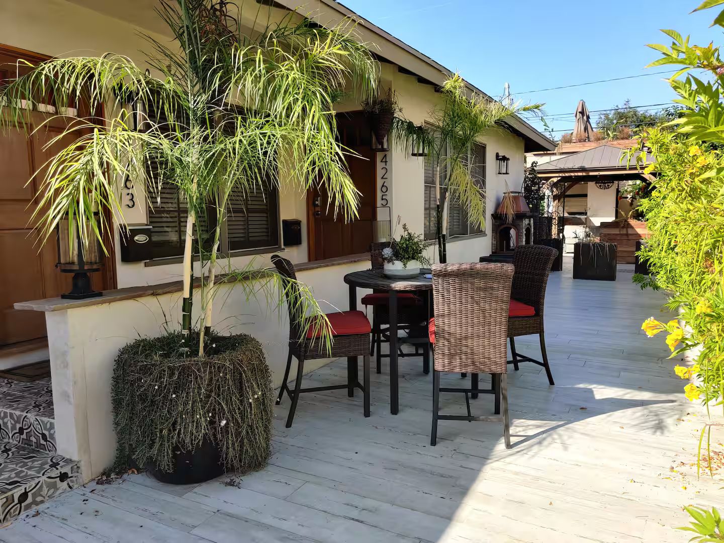 Een terras buiten met rieten meubels, potplanten en een gebouw met genummerde deuren.