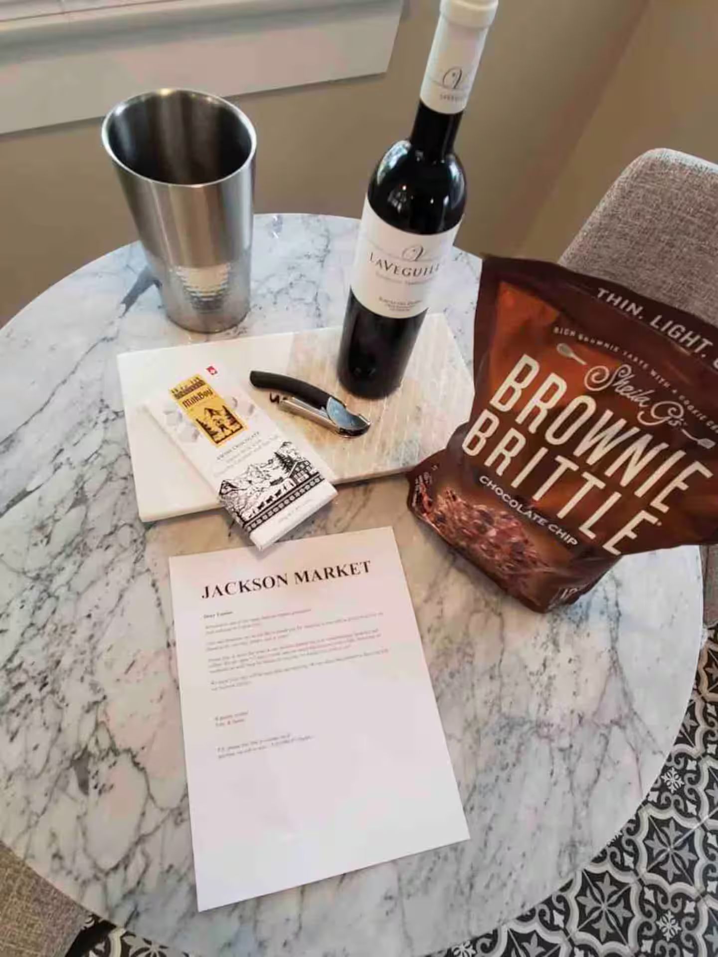 Ett bord av marmor med en vinflaska, en påse med brownies, en penna, ett anteckningsblock och en mugg.