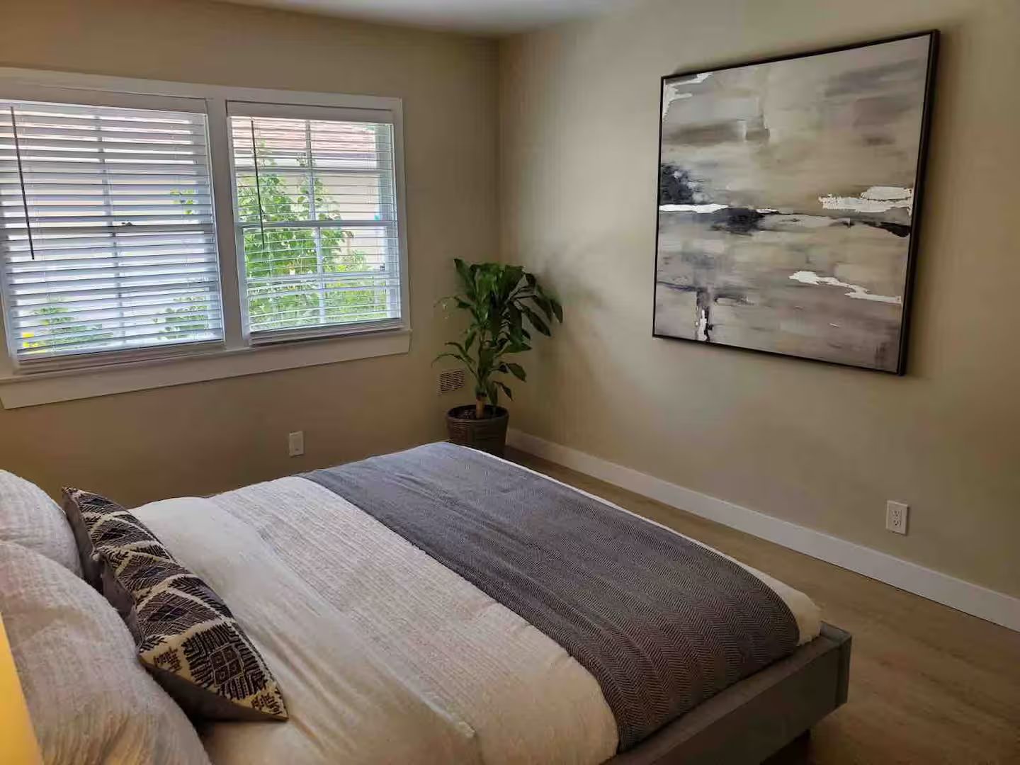 A bedroom with a bed, a window with blinds, a potted plant, and a wall-mounted abstract painting.