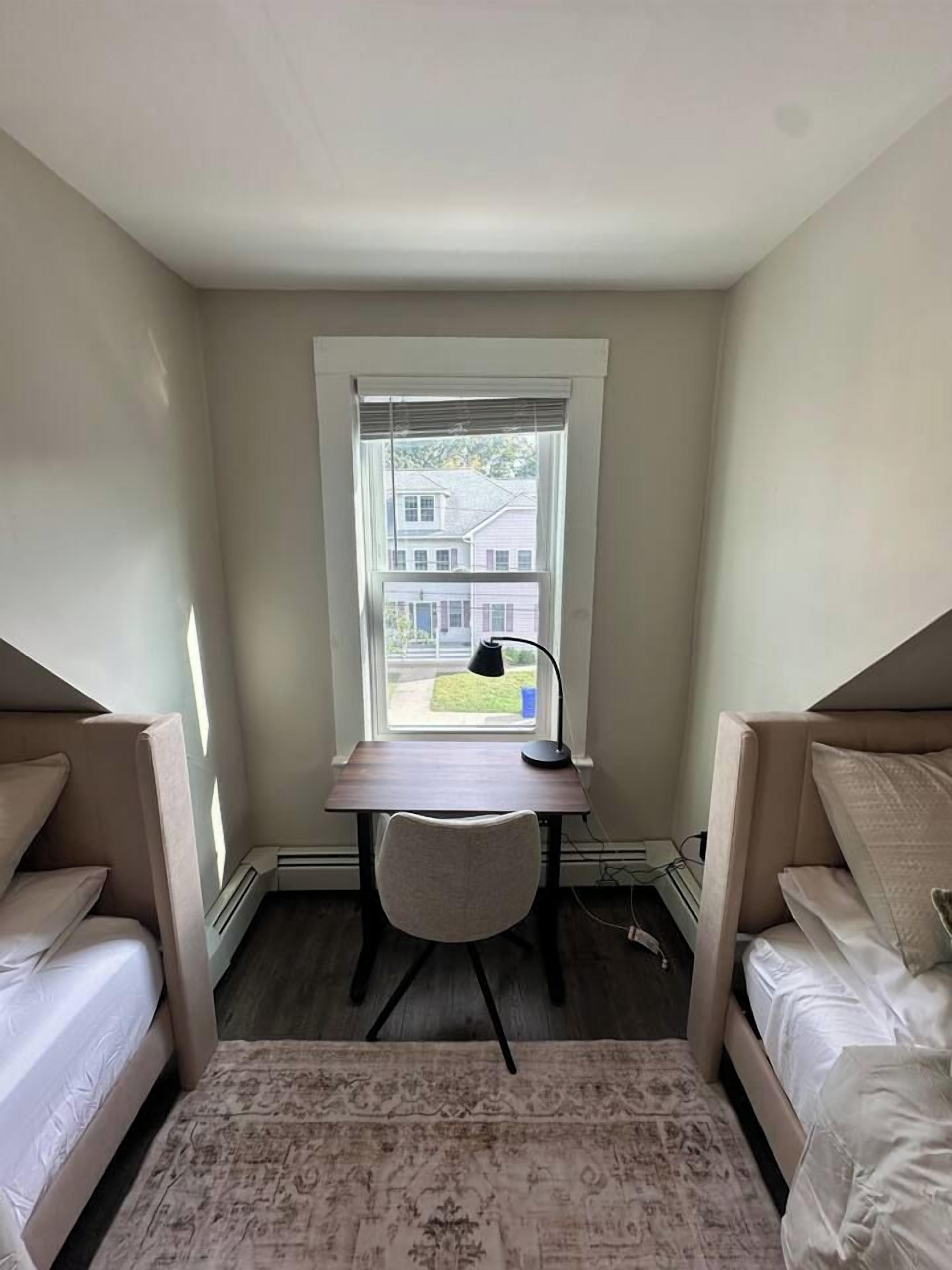 Une chambre avec un bureau et une chaise, une fenêtre avec des stores et une vue sur une maison à l’extérieur.