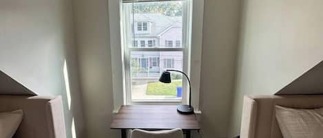 Une chambre avec un bureau et une chaise, une fenêtre avec des stores et une vue sur une maison à l’extérieur.