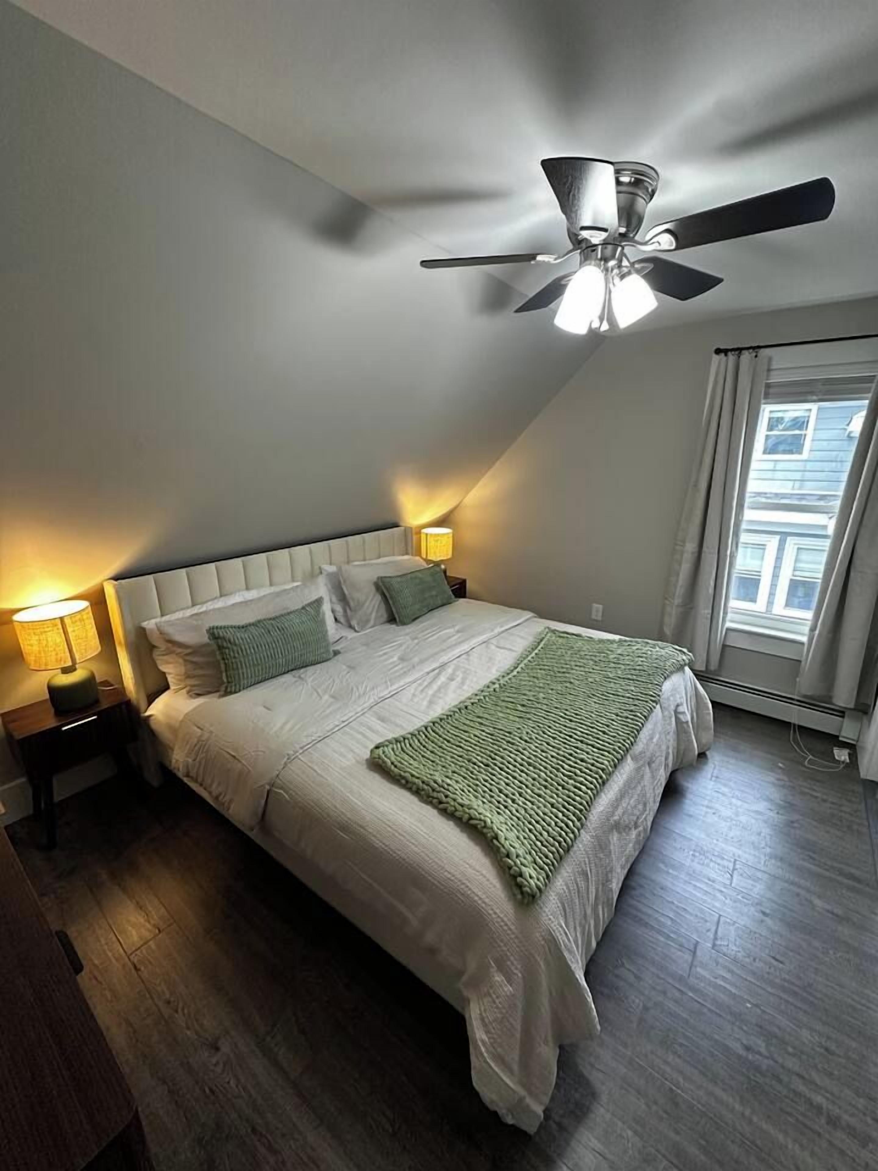 A bedroom with a bed, a ceiling fan, a window, and a nightstand with a lamp.