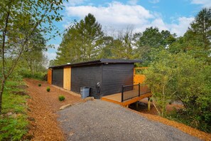 A small, dark-colored building with a wooden deck and a black railing, surrounded by trees and a gravel path. - Elk Stargazer | Roll-Out Bed, Hot Tub & Sauna (Benezette)