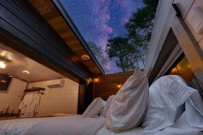Quarto com cama, almofadas, mesa de cabeceira, roupão e janela com vista para o céu noturno.