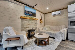 A cozy living room with a stone fireplace, a white sofa, a white armchair, a round coffee table, and a guitar on the wall. - Elk Stargazer | Roll-Out Bed, Hot Tub & Sauna (Benezette)