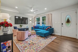 Een moderne woonkamer met een blauwe bank, een roze fauteuil, een televisie en een decoratieve vaas met rode bloemen.