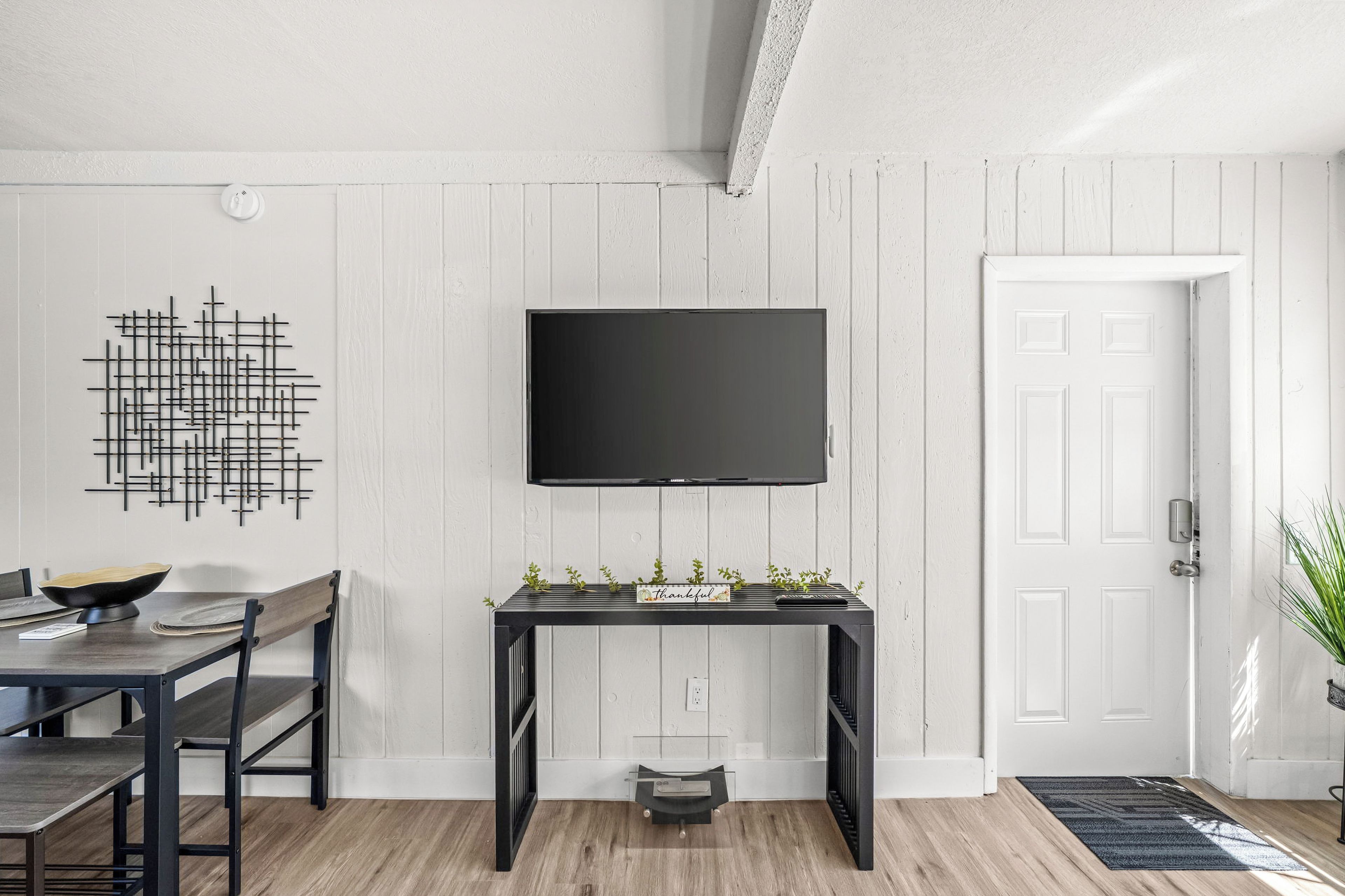 Una sala moderna con comedor, un televisor negro montado en la pared, un adorno en la pared y una puerta con una planta pequeña a su lado.