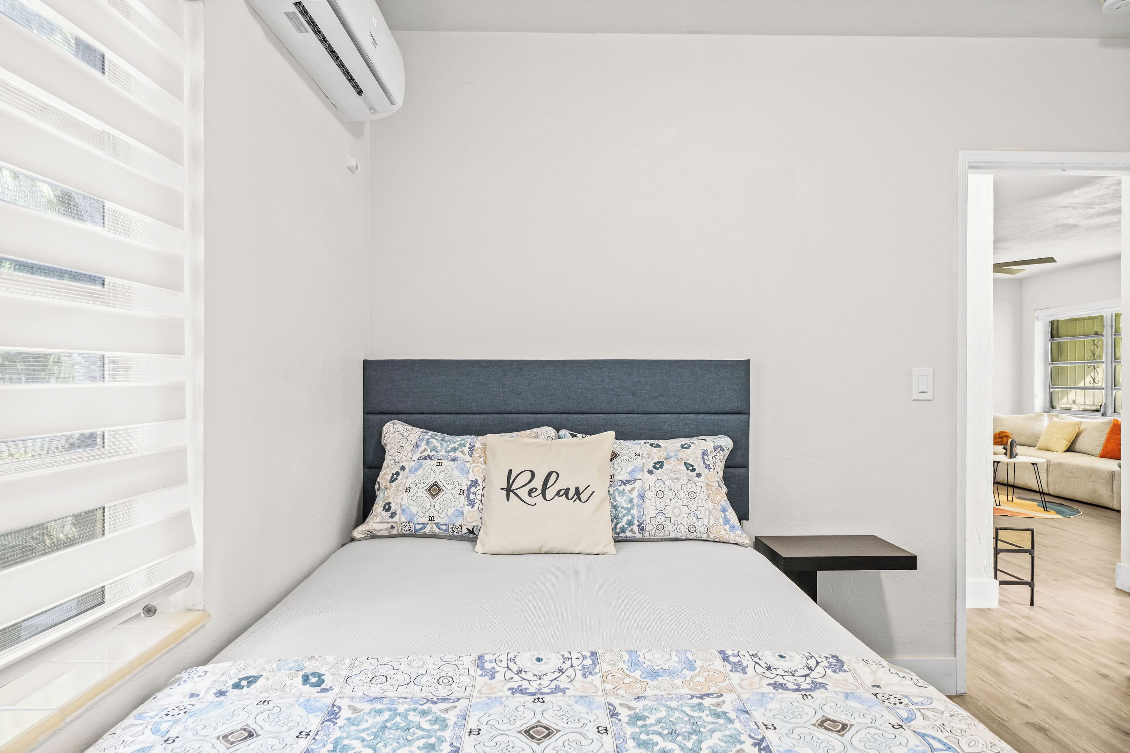 Un dormitorio con cama, una mesita de noche, una ventana con persianas y vistas a otra habitación.