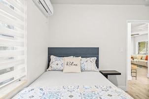 Un dormitorio con cama, una mesita de noche, una ventana con persianas y vistas a otra habitación.