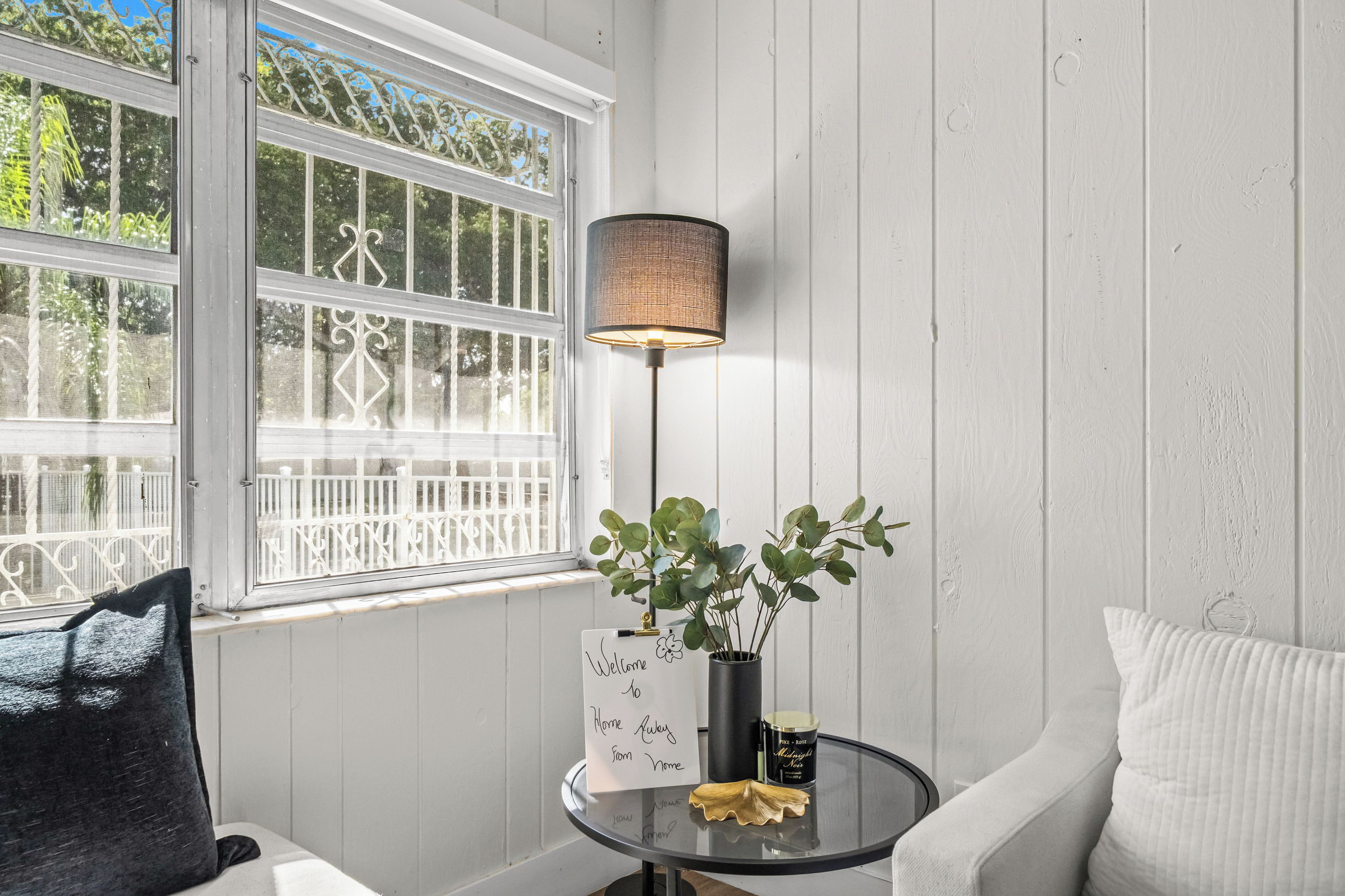 A room with a white wall, a window with a white railing, a black floor lamp, a small table with a plant, a candle, and a welcome sign.