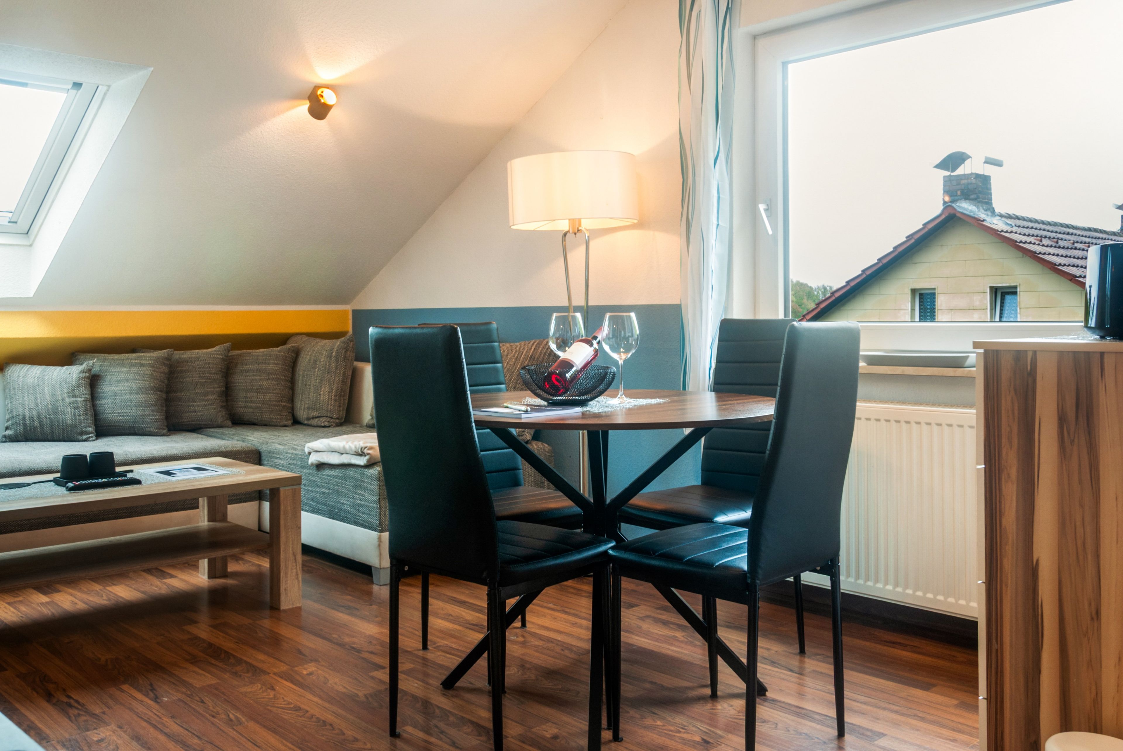 Ein gemütliches, helles Wohnzimmer mit Essbereich, einer Couch und Blick auf ein Haus durch ein Fenster.