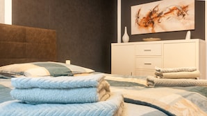 A neatly made bed with folded towels, a white dresser with drawers, and a painting on the wall.