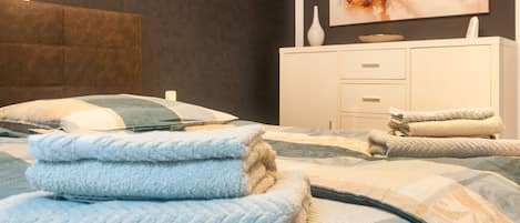 Ein ordentlich gemachtes Bett mit gefalteten Handtüchern, eine weiße Kommode mit Schubladen und ein Bild an der Wand.