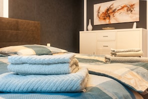 A neatly made bed with folded towels, a white dresser with drawers, and a painting on the wall.