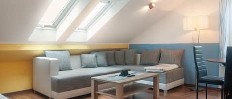 Une chambre moderne de style loft, avec un plafond en pente, un ensemble de canapés, une table basse, une lampe et une vue sur l’extérieur grâce à des fenêtres de toit.