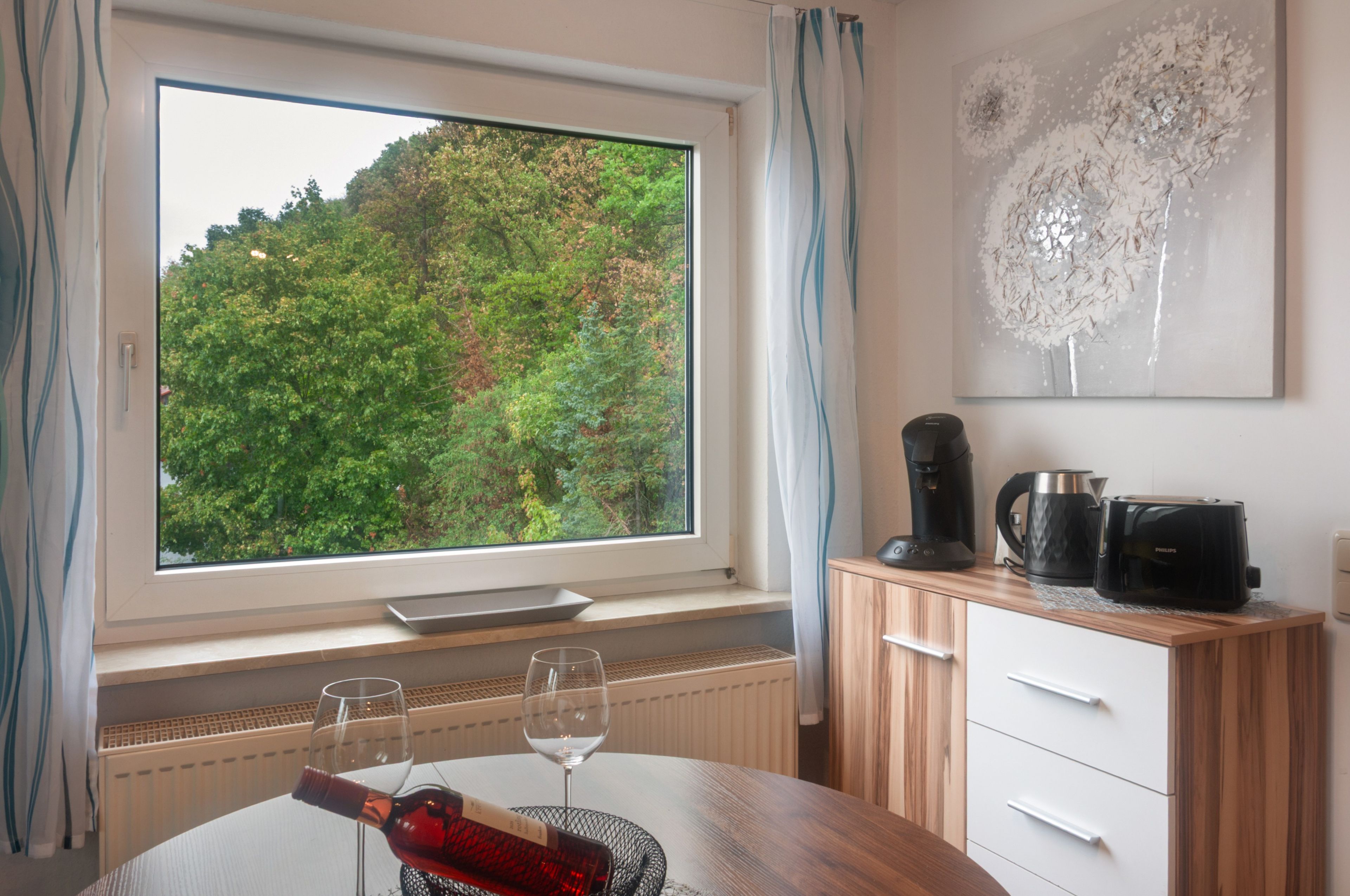 A room with a large window, a dining table with two wine glasses and a bottle, a coffee maker, and a kettle on the counter.