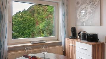A room with a large window, a dining table with two wine glasses and a bottle, a coffee maker, and a kettle on the counter.
