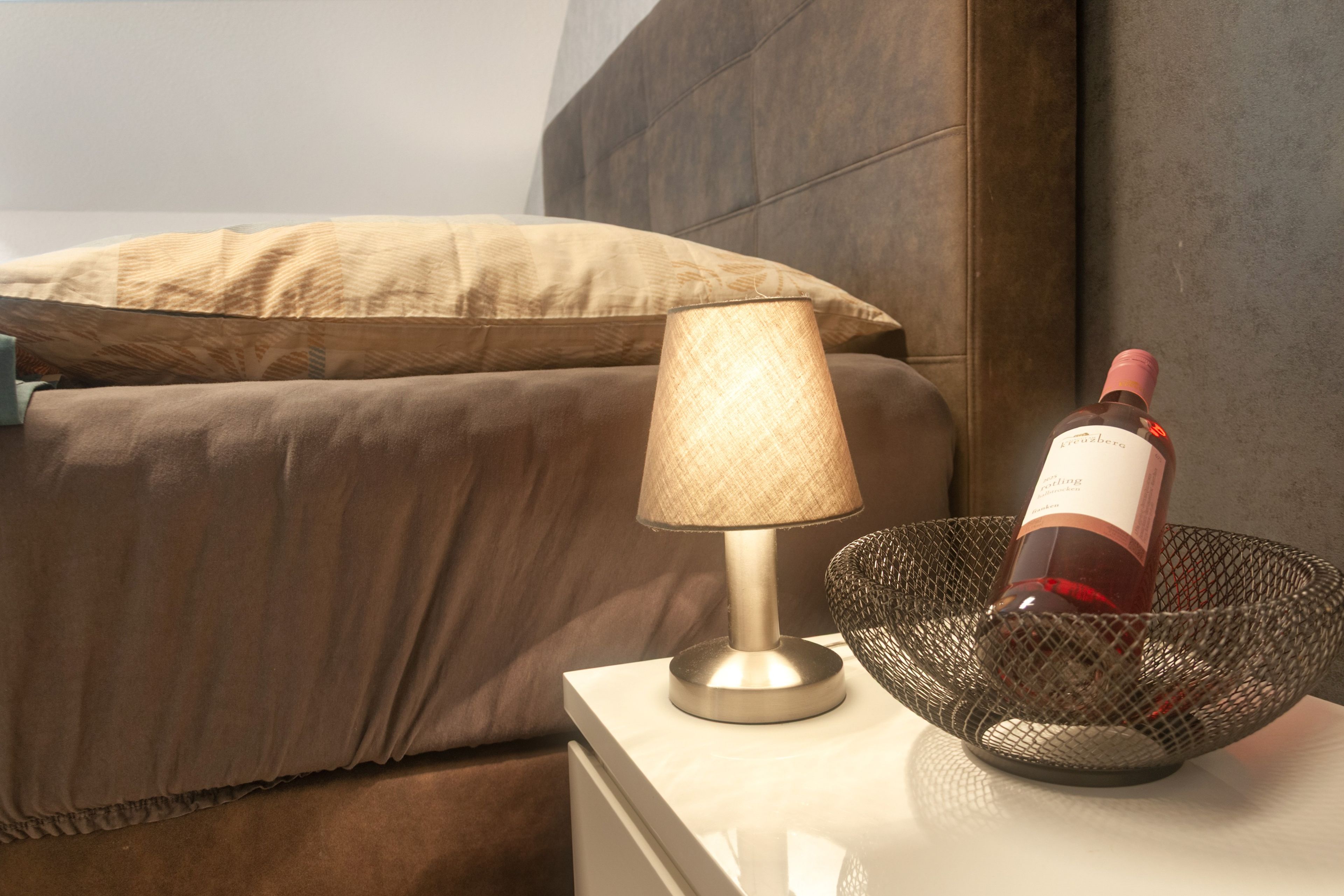 A bed with a headboard, a bedside lamp, and a wine bottle in a wire basket on a nightstand.
