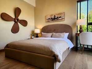Une chambre avec un grand lit, un ventilateur fixé au mur, un tableau, une lampe de chevet et une vue par la fenêtre.