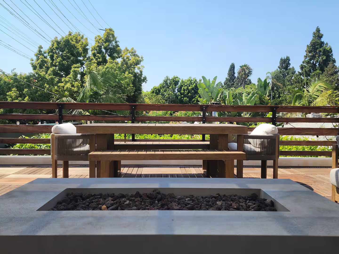 A wooden bench with cushions, a concrete planter with rocks, and a view of greenery and trees.
