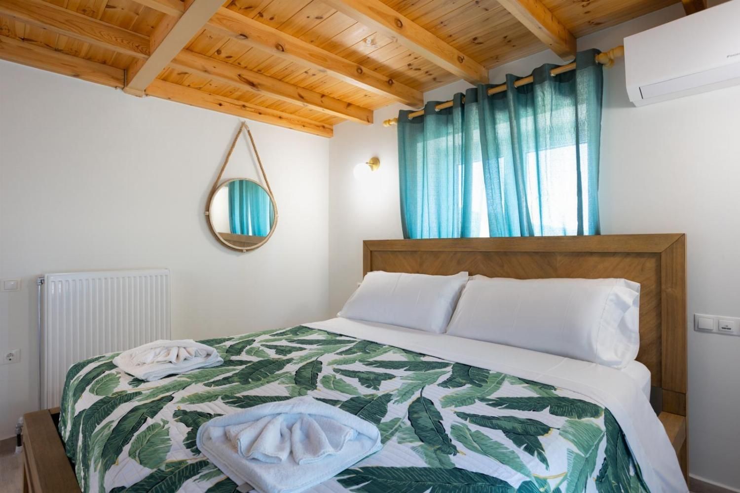 A hotel room with a wooden ceiling, a bed with a leaf-patterned bedspread, a round mirror, and a window with blue curtains.