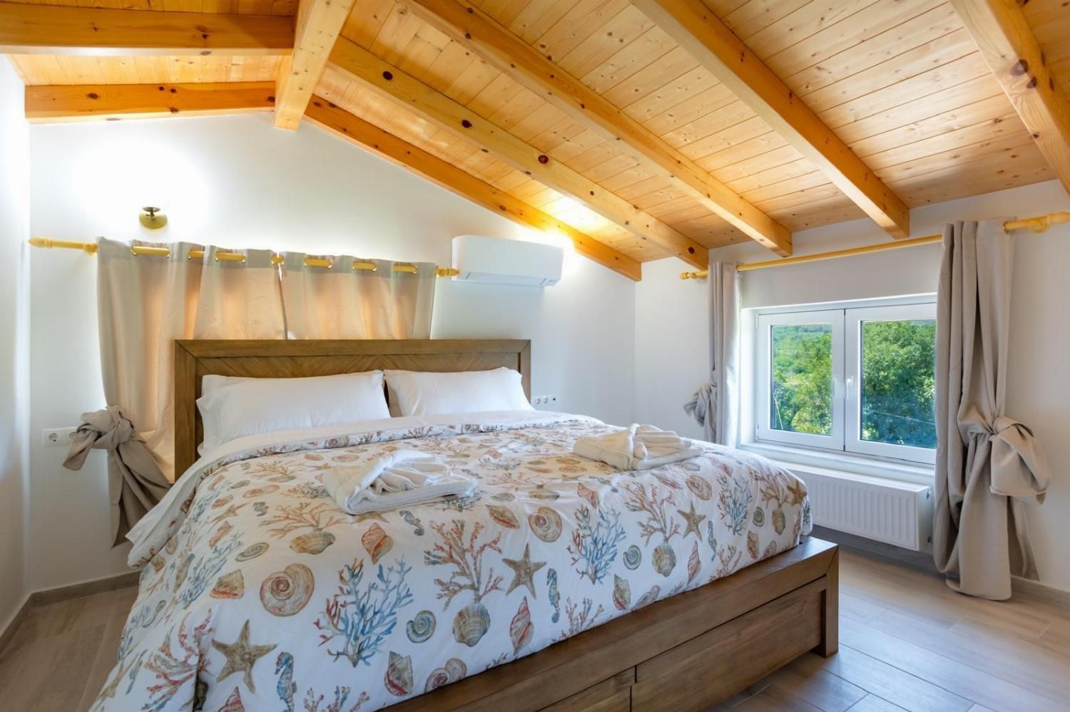 Une chambre avec un plafond en bois, un lit recouvert d’une couvre-lit à motifs floraux, une fenêtre donnant sur la verdure et un radiateur placé sous la fenêtre.