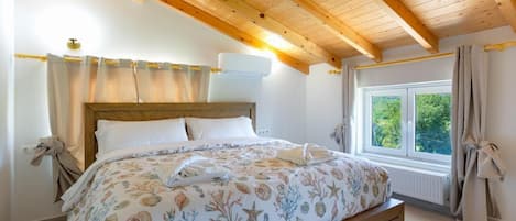 A bedroom with a wooden ceiling, a bed with a floral bedspread, a window with a view of greenery, and a radiator under the window.