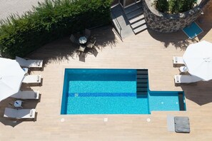 Eine Vogelperspektive auf einen rechteckigen Pool mit Steinrand, umgeben von Liegestühlen und Sonnenschirmen, sowie einem kleinen Tisch mit Stühlen.