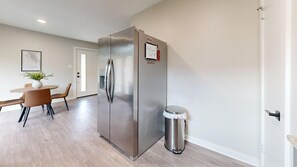 A modern kitchen with a stainless steel refrigerator, a trash bin, and a dining area with a table and chairs. - *NEW* Hullabaloo Hangout - 1.1 miles to Kyle Field! (College Station)