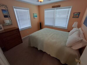 A bedroom with two windows, a bed, a dresser, and a wall-mounted heater. - Canal Front Home on sought out street - Unique, Cozy and Ready for YOU! (Ocean Isle Beach)