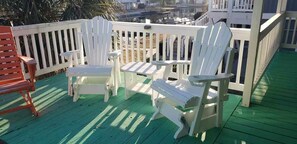 En terrass med vita Adirondack-stolar och en röd stol, grönt trädäck och ett vitt räcke.